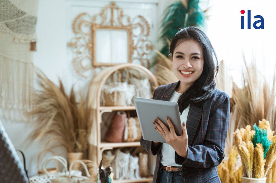 Một người đàn ông đang ngồi trên ghế bành, dùng laptop và có một chiếc điện thoại đặt trên bàn, tượng trưng cho việc quản lý tài chính